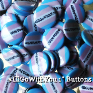 Image Description: Pile of small circular buttons featuring the #IllGoWithYou logo, which is black serifed text reading "#IllGoWithYou" centered on the button over the transgender pride flag, which is a light blue bar on top, followed by a light pink bar, a white bar in the middle, followed by another light pink bar, then another light blue bar at the bottom. Superimposed over the bottom right side of the picture is white serifed text reading "#IllGoWithYou 1" Buttons"